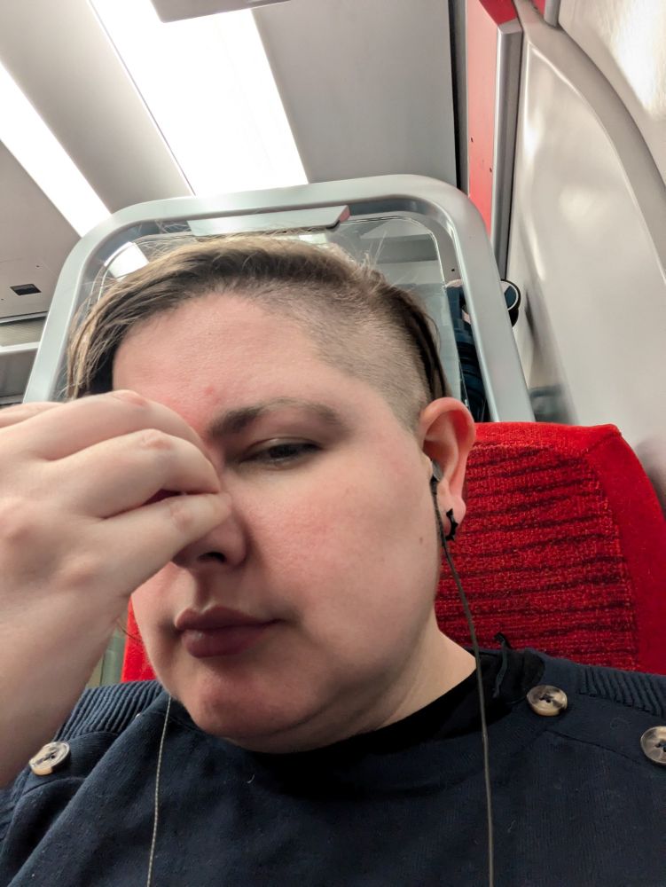 A picture of a white person at the end of their tether, sitting on a train (red seat). I'm wearing a navy jumper with tortoiseshell buttons on the shoulders, and my hair is shaved on the side with the rest falling over the opposite part of my face. I have headphones in and a black spiky earring