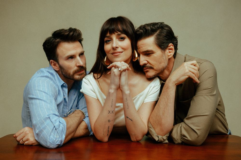 Chris Evans, Dakota Johnson, and Pedro Pascal in promotion photos for the upcoming movie The Materialists. Dakota is in the middle looking straight ahead with Chris and Pedro leaning on her and looking at her. 