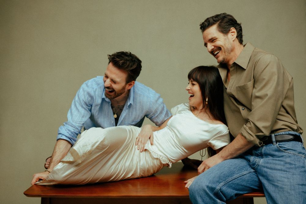 Chris Evans, Dakota Johnson, and Pedro Pascal in promotion photos for the upcoming movie The Materialists. Dakota is laying on a table leaning against Pedro with Chris leaning against her. All three appear to be laughing.