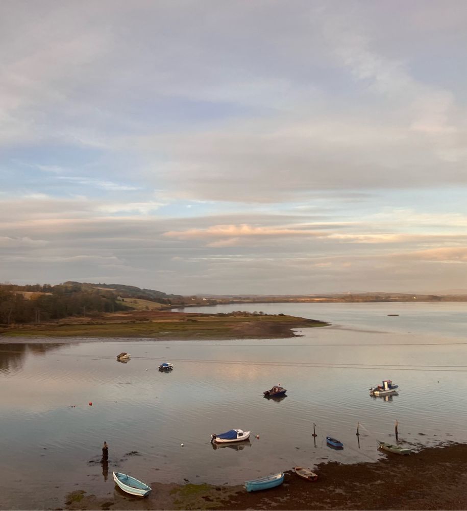 Pastel colors of sunrise filter through clouds above a lake. Small boats are scattered in the water and on the muddy shore. 