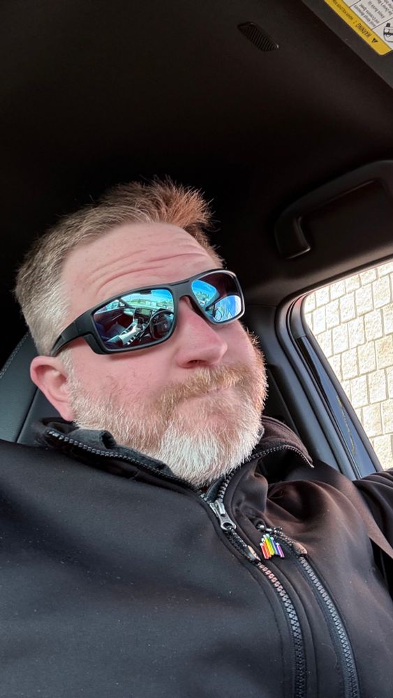 A very white dude in a cliched “white guy in car” selfie down to the shiny sunglasses.

A Star Trek IDIC rainbow pin breaks up the monotone black colors he’s otherwise wearing. 