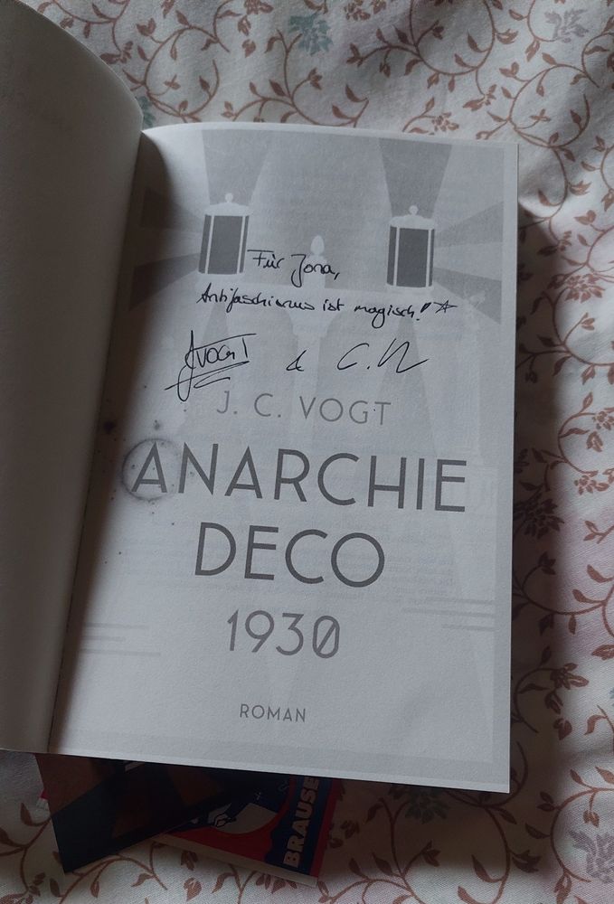 Das aufgeschlagene Buch "Anarchie Deco 1930" von J. C. Vogt liegt auf einer geblümten Decke, über dem Titel steht "Für Jona, Antifaschismus ist magisch", darunter haben die Autor*innen unterschrieben