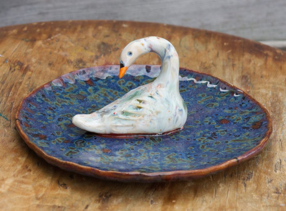 A swan glazed with a rainbow effect (using chips of glaze) sits in the middle of a plate. The surface of the plate is dark and mysterious and kind of algae-looking, in shades of dark blue, green and redbrown