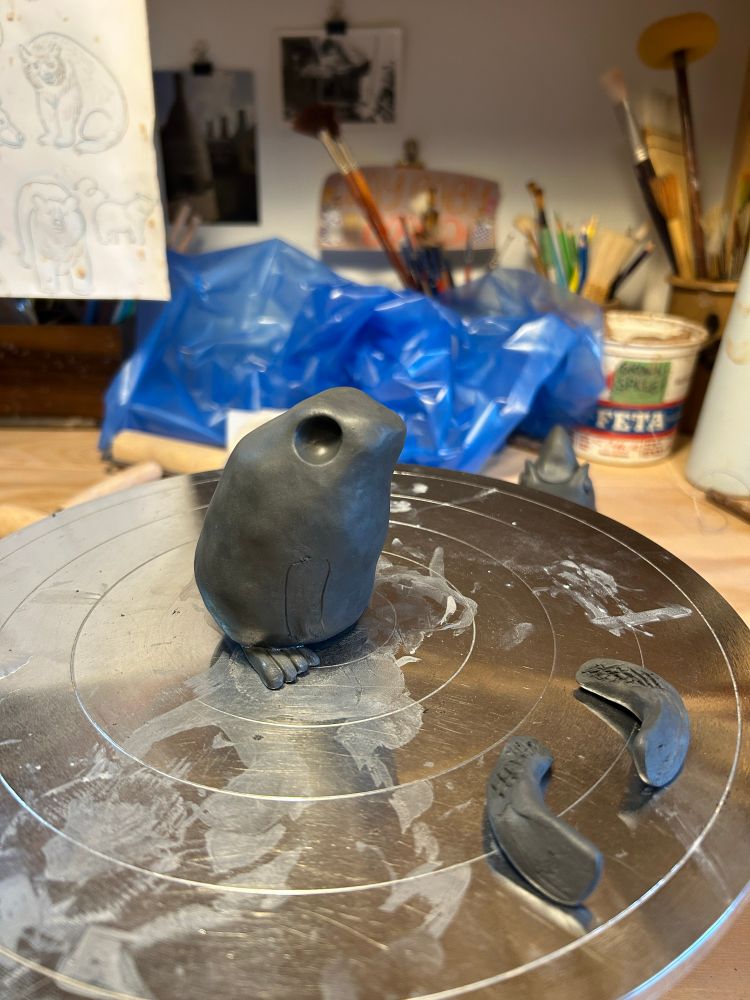An in-progress toad made from black clay sits on a steel banding wheel. Its future arms are lying to the right of it.