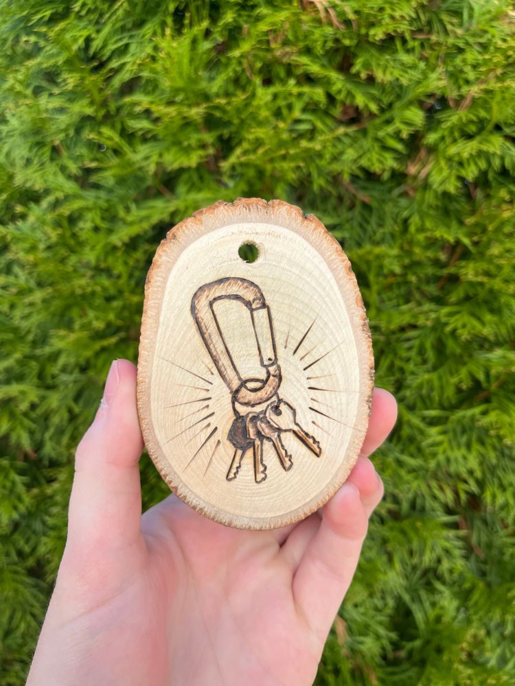 Hand holding a small wooden cross-cut of a tree with a ring of keys image burned into it. 