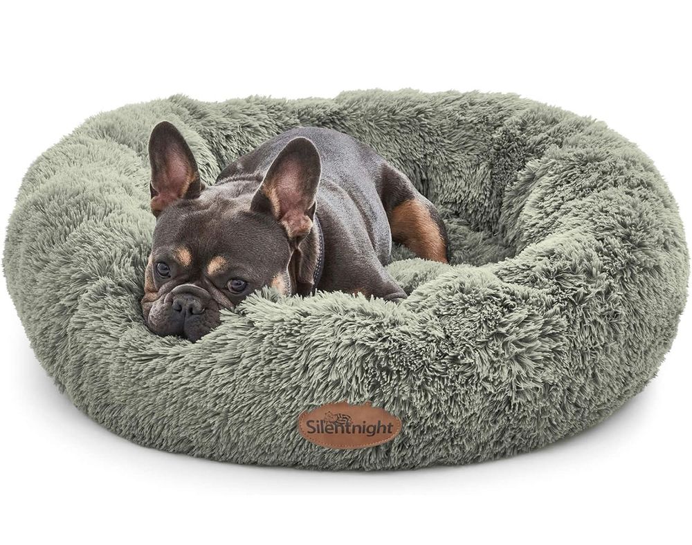 A sage green fluffy round circular dog bed, with the label Silent Night on. There is a small Boston terrier snoozing in it, who has tan and black markings