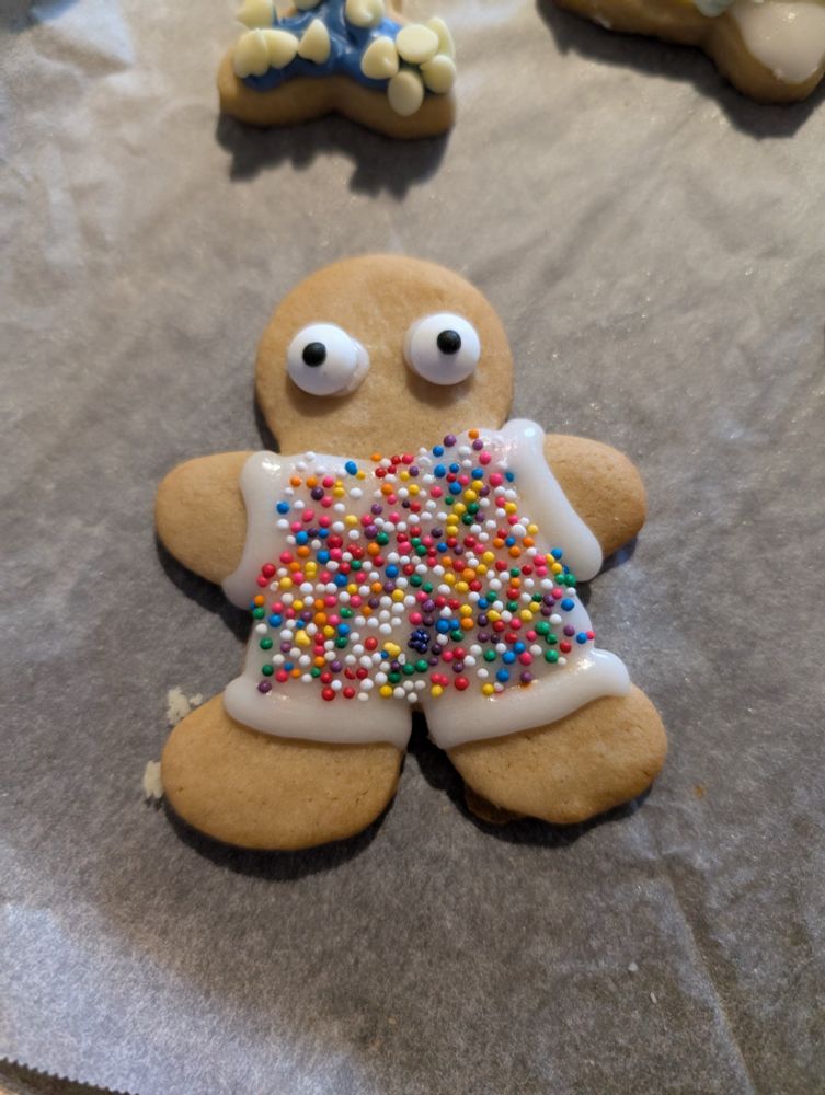 A person-shaped brown sugar cookie with wide, terrified eyes and a rainbow sprinkle jumpsuit.