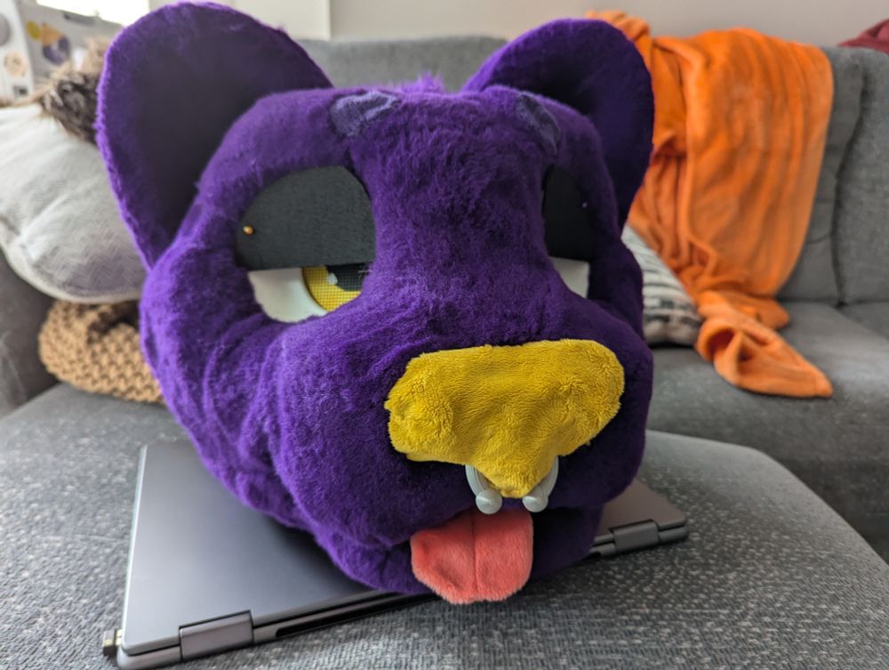 Purple cat fursuit head with yellow eyes yellow nose, and silver septum piercing.