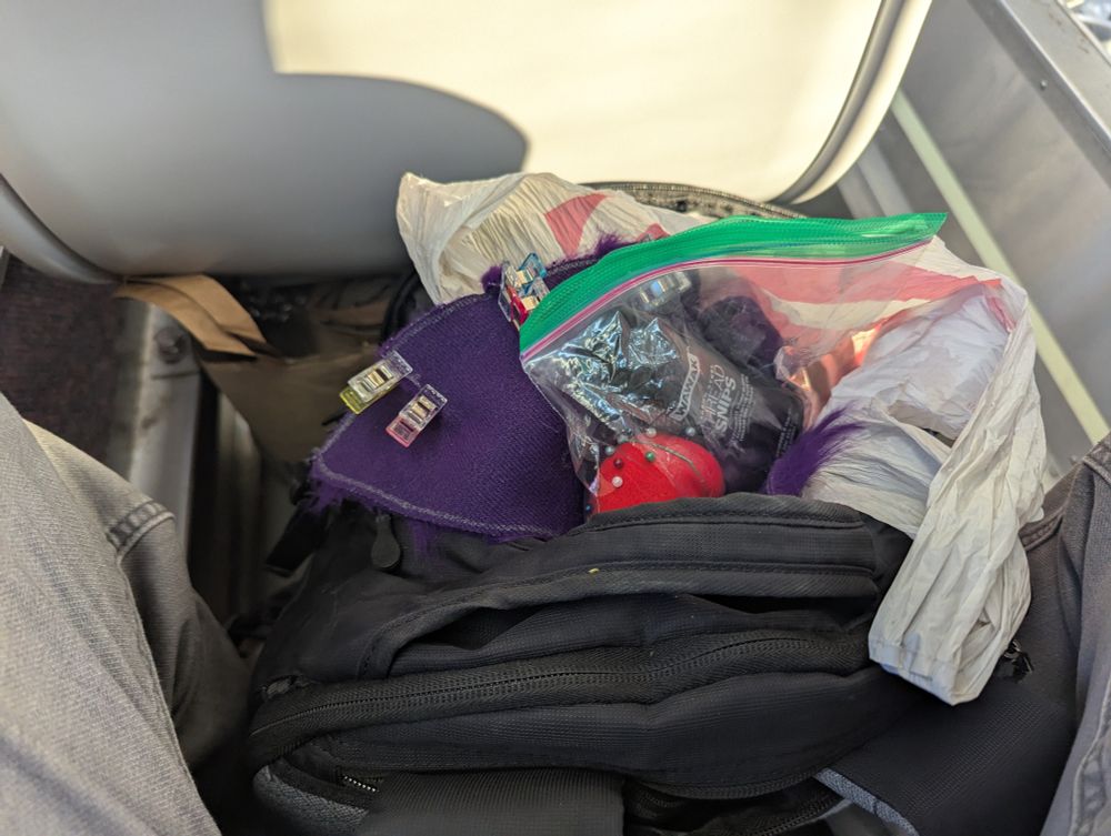 backpack sitting between my legs and behind a train seat, with fur and sewing supplies on top of the bag
