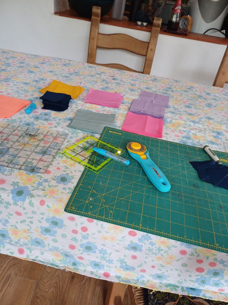 Coloured squares (pink, purple, yellow and orange), a cutting mat and quilting squares all laid out on a very 1970s tablecloth from my mother 