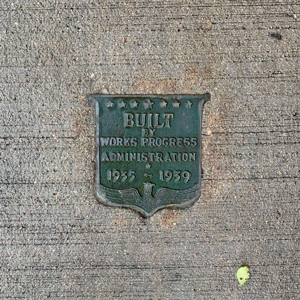 brass marker set in concrete, shaped like a shield with stars at the top and an eagle at the bottom, with the words: Built by Works Progress Administration, 1935–1939