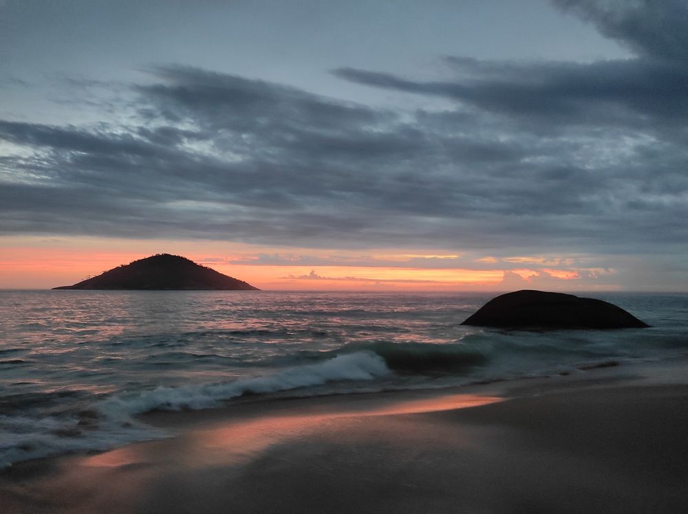 Uma linda praia ao pôr-do-sol, com os tons laranjas e azuis acinzentados do céu se refletindo no mar e na faixa onde o mar encontra a areia