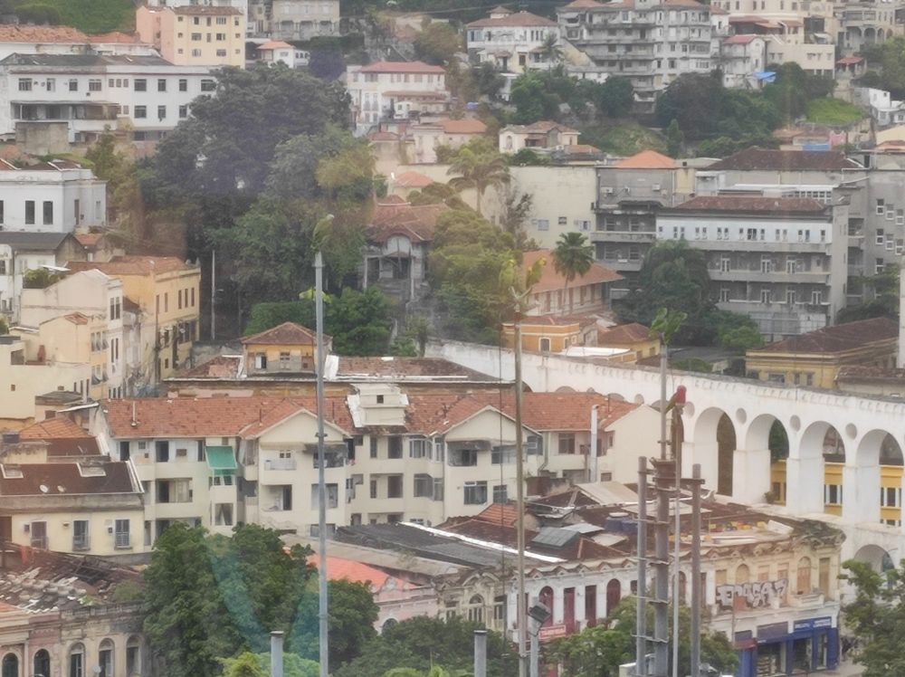 Foto dos Arcos da Lapa (RJ) ao fundo, vistos de cima, com papagaios/maritacas apoiadas sobre antenas em um prédio no plano médio.