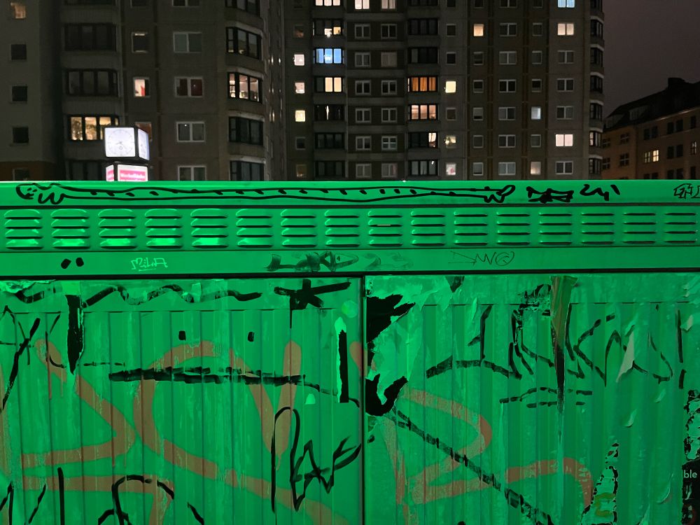 A sidewalk electrical box, painted light gray, with graffiti scrawled here and there. Along the top edge, a simple cartoon of a smiling animal; it is probably 50 cm/20 in long and about 2 cm/1 in tall. It might be a wiener dog. It has spots along its back. In the background: an apartment building, several windows lit from inside. 