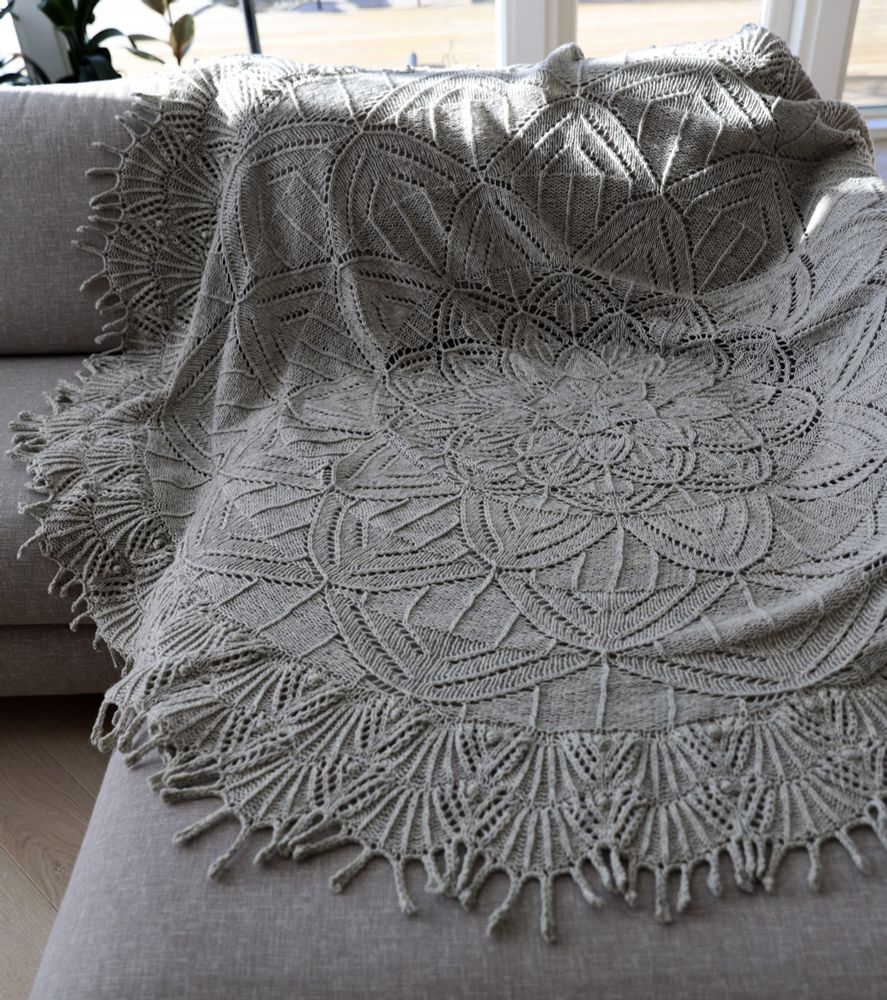 A light grey, round handknit lace blanket on a grey couch.