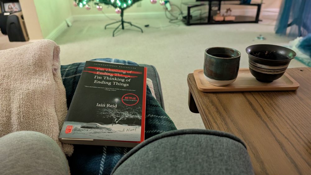 Ben is sitting on a couch with the book "I'm thinking of ending things" resting on top of some blankets in front of him. There's a teacup full of a black tea called "Qing Xin Red" and a gaiwan beside it full of the tea leaves. It is moody and warm, and there's light from the christmas decorations nearby.
