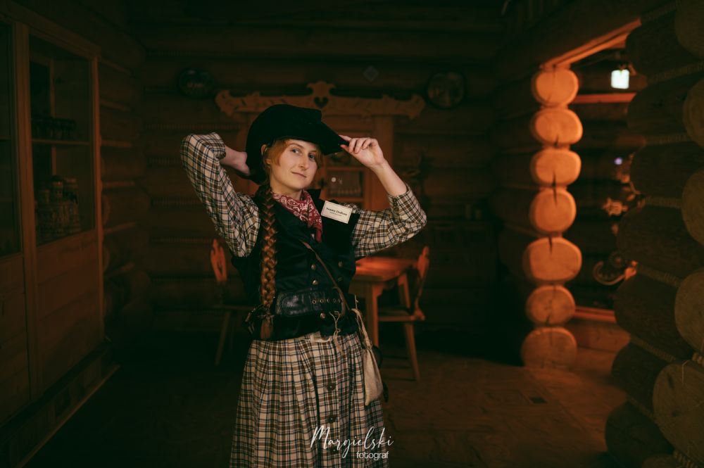 A portrait photo of a wild west character in a checkered dress and a black waistcoat, with black cowboy hat and a long red braid. The background is of a room in a wooden restaurant. 