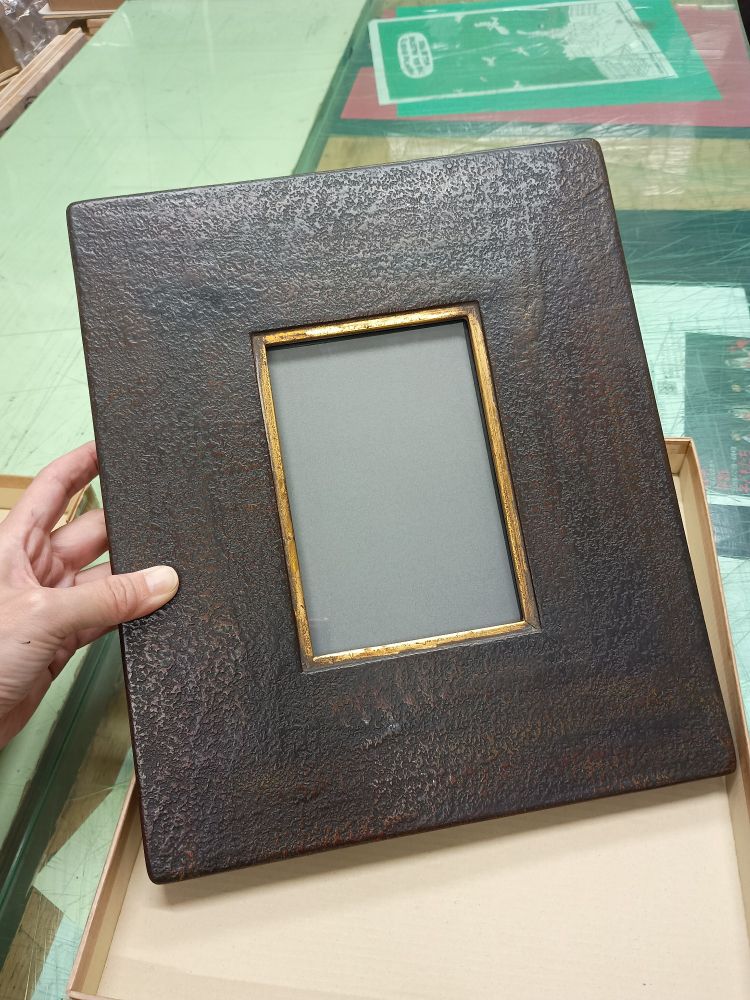 はがきサイズの作品が入る額縁。70㎜くらいの巾の平らな板のような額で、作品の周りに面金がされています。ザラザラとした質感で暗い茶のような色に塗られています。左手で少し立てて撮った写真です。