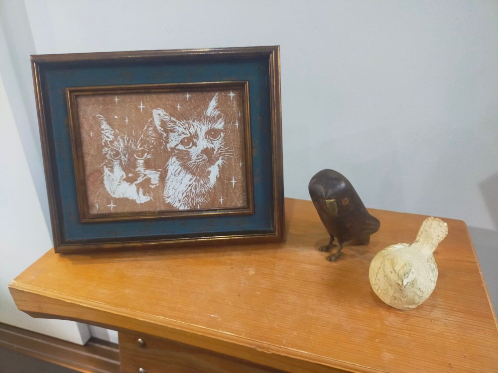 会場入口の台に飾られた、額装された作品。
２匹の猫ちゃん達が茶色で刷られています。
額の隣には２羽の小鳥の置物。
猫ちゃん達が狙いを定めている！？