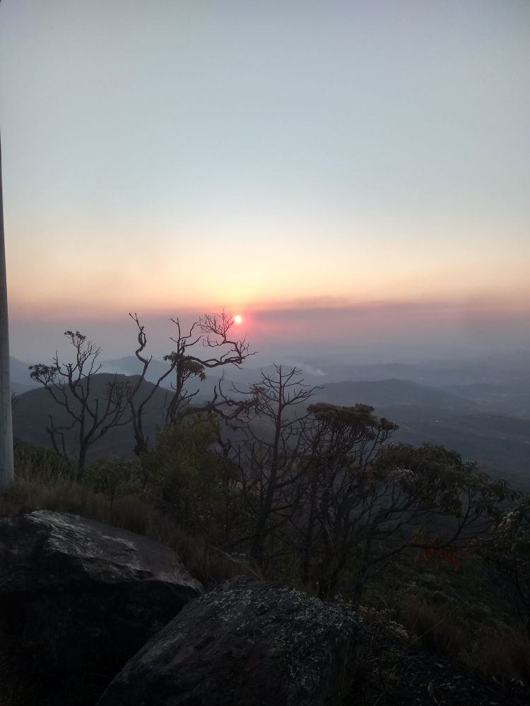 Foto da vista da parte de cima da Serra da Piedade, a vista consiste em várias serras ao fundo, algumas árvores retorcidas e secas em primeiro plano, sol em cor avermelhado e um pouco abaixo do sol um foco de incêndio, o fundo da imagem está um pouco embasado devido a presença de fumaça.