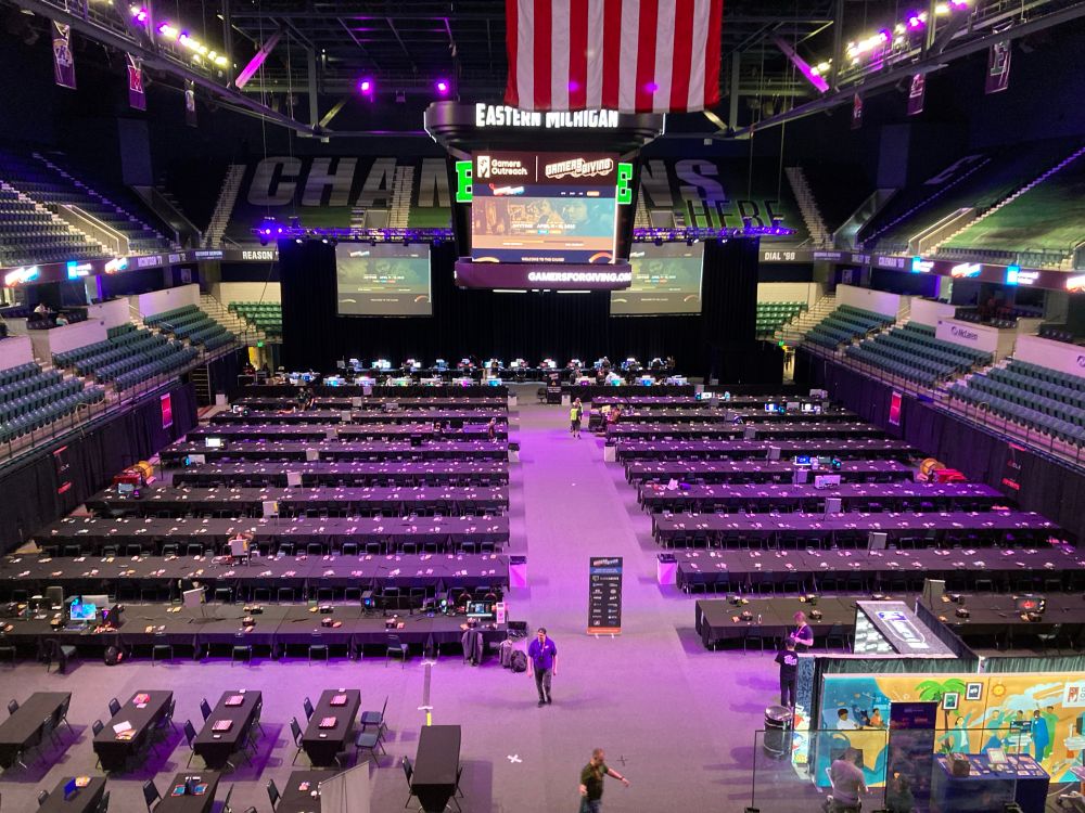 an arena full of tables under white and pink lighting. a few people are visible talking or working.