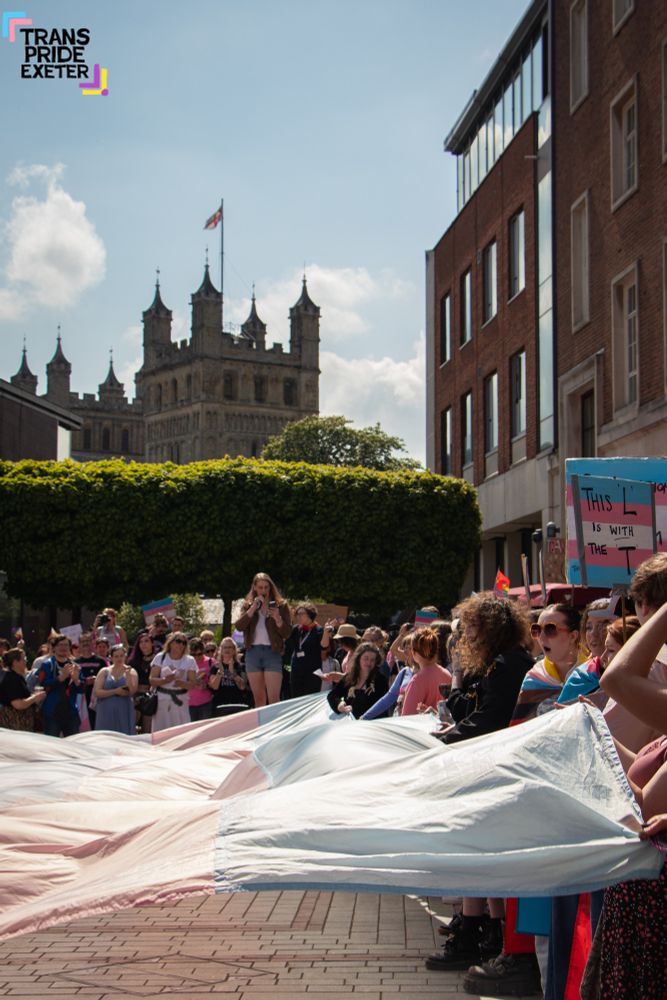 A crowd gather around a huge trans flag, while a speach is being made