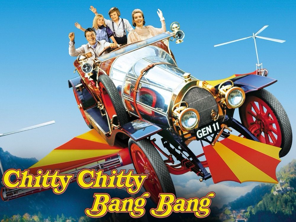 Promotional image of the movie Chitty Chitty Bang Bang featuring the iconic flying car with red-spoked wheels, shiny chrome accents, and yellow-and-red striped wings extended from the sides. The license plate reads “GEN 11.” Seated in the car are the main characters: inventor Caractacus Potts (played by Dick Van Dyke), his two children Jeremy and Jemima, and Truly Scrumptious. They are smiling and waving as the car soars through a bright blue sky, with propellers spinning above the rear wings. In the background, lush green hills and quaint European-style villages are visible far below. The movie title, “Chitty Chitty Bang Bang,” is displayed in bold yellow and red letters at the bottom.