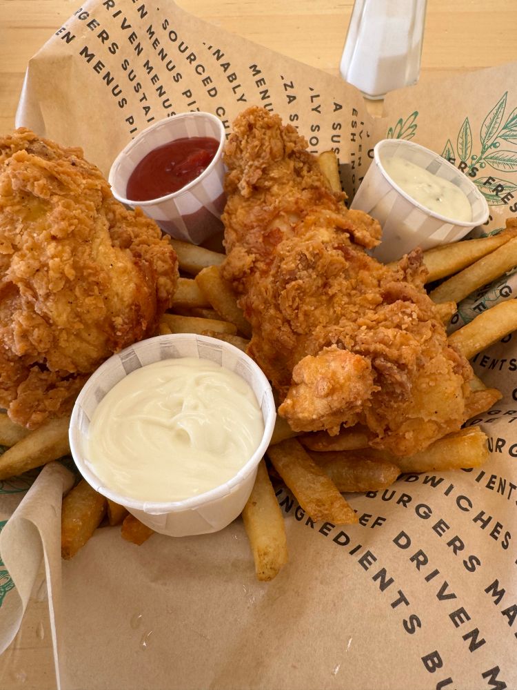 A plate of crispy fried chicken tenders served on a bed of golden French fries. The meal is accompanied by three small paper cups of dipping sauces: ketchup, ranch dressing, and a creamy white sauce, possibly mayonnaise or garlic aioli. The food is arranged on branded brown parchment paper with printed text emphasizing sustainability and fresh ingredients. In the background, a white salt shaker is visible on the light wooden table. The presentation is casual and appetizing, with a focus on comfort food classics.