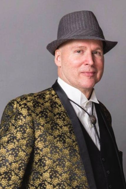 a pale, middle-aged bald man wears a gray pinstripe fedora, gaudy gold blazer, dark gray undervest, frilly white collar dress shirt, a bolo tie, and a gaudy floral print gold jacket. he is barely smiling.