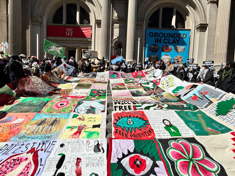 steps at the entrance of the of the Met Museum covered in massive quilt made up of artwork, mostly in reds, pinks, greens, black and white, expressing solidarity with Palestine. Images include a pomegranate, a flower made of watermelon slices, poppies breaking through the apartheid wall, an eye with a poppy against a green and white checkered background. Photo by Alexa B Wilkinson.