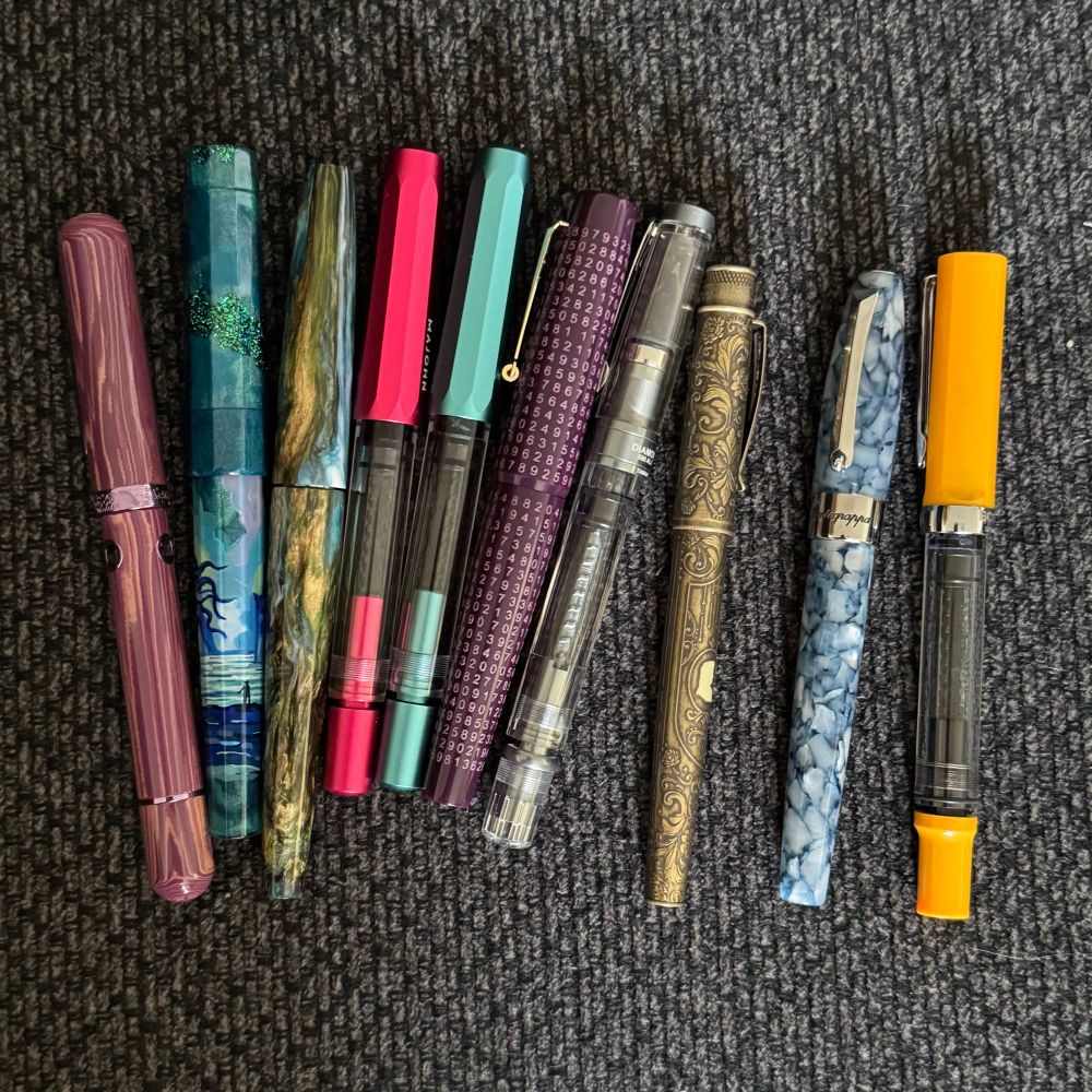 Ten fountain pens of various brands, styles and colors, arrayed on a charcoal gray couch cushion.