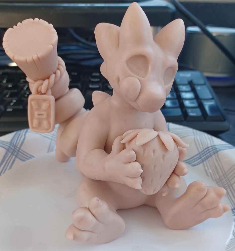WIP sculpt of an anthro gecko with a string bead tail, she is holding a strawberry in her arms and is licking her mouth looking down at it hungrily.