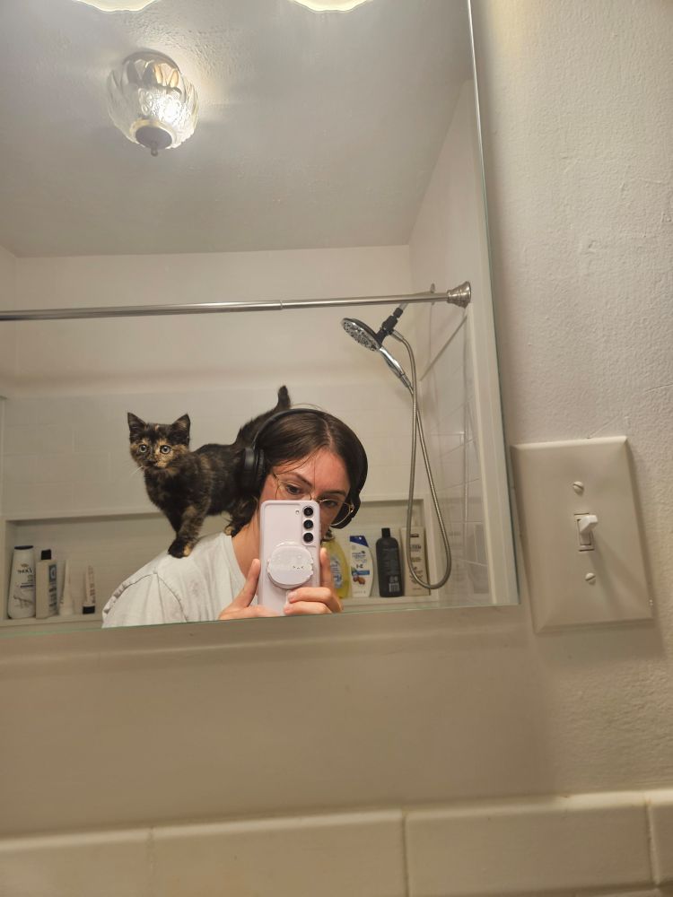 Mirror selfie of me (brown hair, headphones, glasses) with Ava (tortoiseshell kitten) standing on my shoulder 