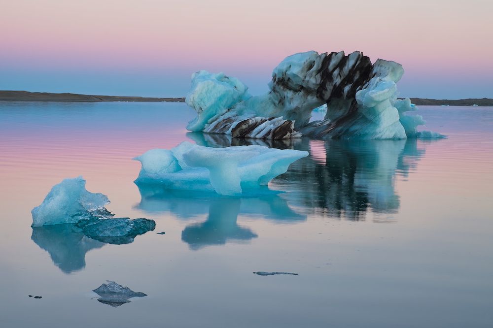 An iceberg with visible strata of volcanic ash deposits floats in the glacial lagoon of Jokulsarlon in south Iceland, under the pink dawn-like glow of the midnight sun.