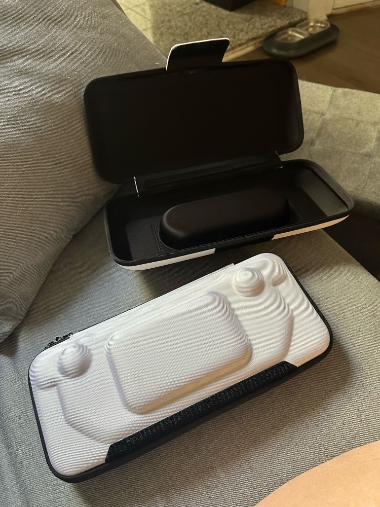 a closed white slim steam deck case sits next to an open larger protective steam deck case on a couch. the slim case perfectly fits inside of the larger case. 
