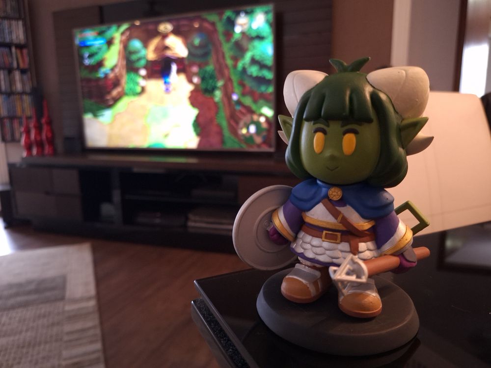 Fotografia de uma estatueta de tiefling de cores verde e vestes de clériga em cima de uma mesa no ambiente de uma sala de estar. Uma TV desfocada no fundo reproduz o jogo "The Legend of Zelda: Echoes of Wisdom", o formato da estatueta está dentro do estilo artístico do respectivo jogo.