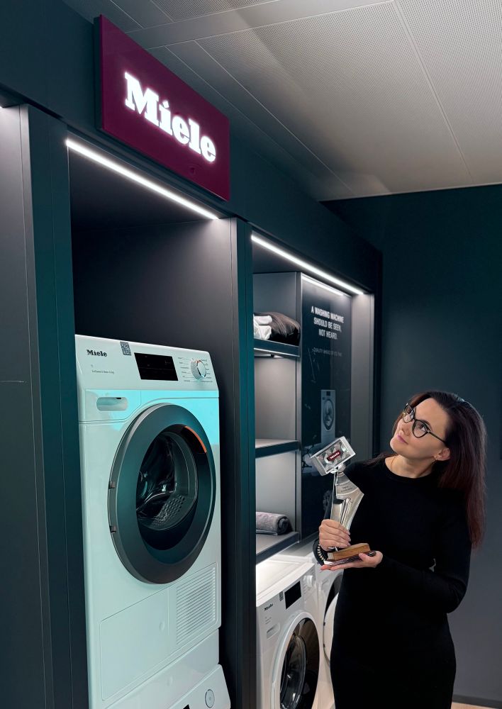 A woman holding a trophy and looking at a Miele washing machine in a showroom, with the illuminated Miele logo above.