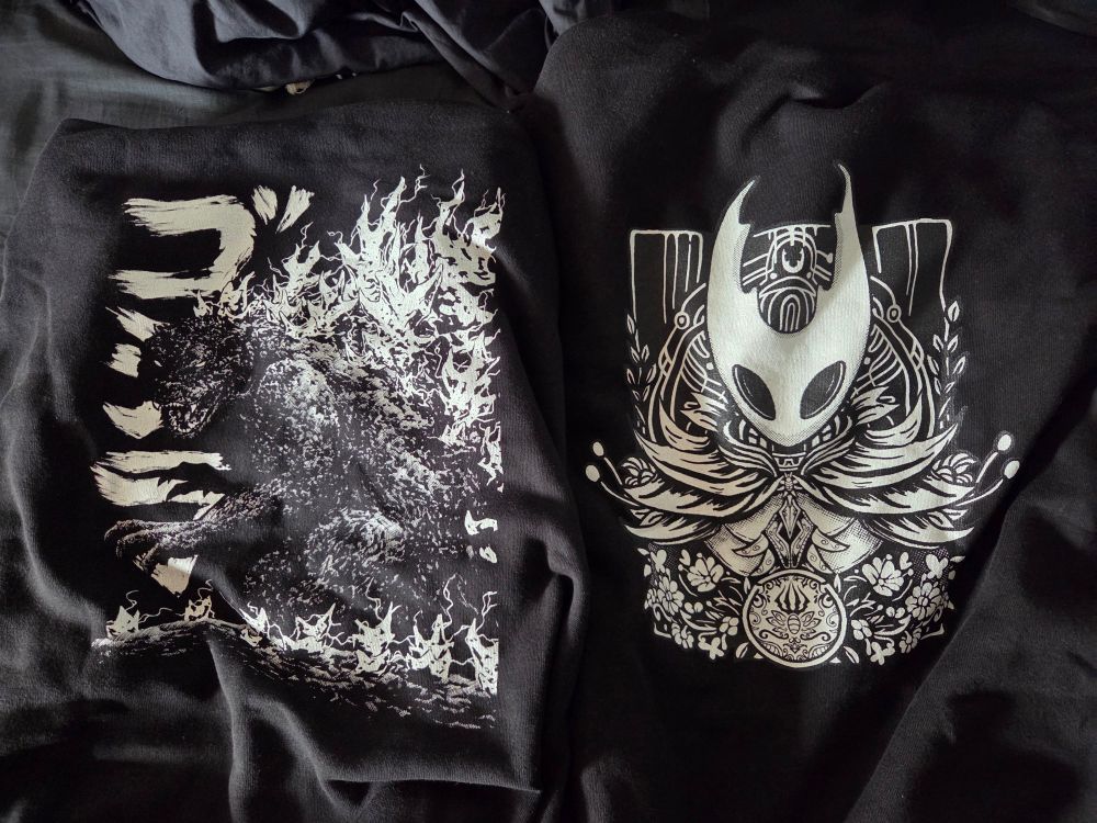 The back of 2 black hoodies showing a black and white image of Godzilla on one and the black and white image of Hornet, from Silksong, on the other.