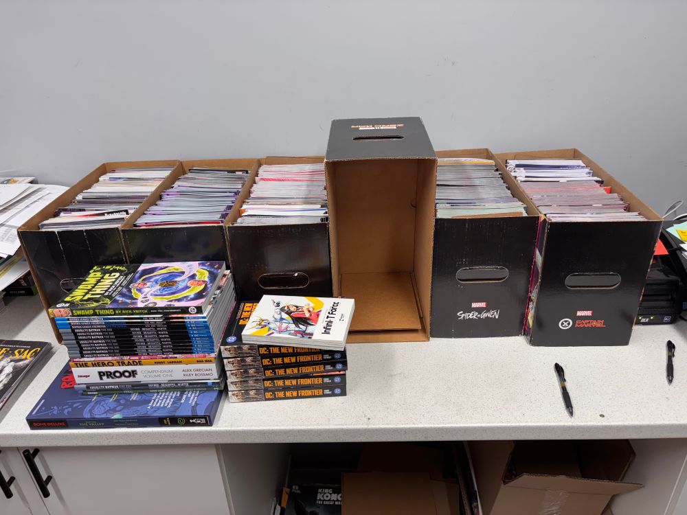 Five short boxes filled with comics and two small piles of books are arranged on a table. Splitting the five boxes is another empty short box - this is where all the file pulls will go until it is full - and then then I’ll grab another box to fill.