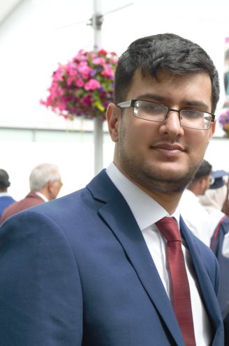 Brown male wearing glasses, short hair and a blue suit with a red tie looking directly at the camera