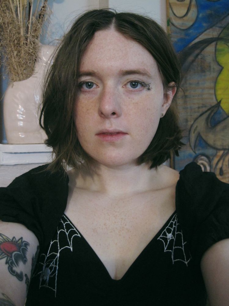 Selfie of Nessie, and a homemade embroidered spiderweb dress with spiderweb eyeliner featuring a little drawn spider hanging from a thread of its silk. She is sitting in front of a cat vase and painting that soup made.
