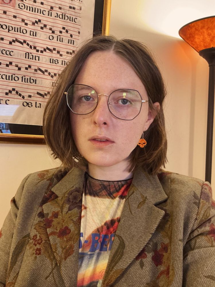 Recent photo of Nessie, standing in front of a folio of Gregorian Chant with a floral blazer and clown shirt on sporting wide rim, glasses, and crocheted orange skull earrings