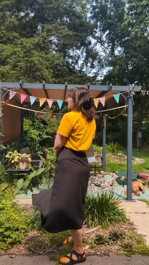 person standing by a messy garden showing off a mid length black skirt. Wearing a yellow shirt and socks and sandals