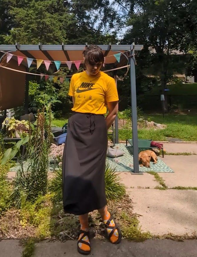 Person standing in front of their messy garden bed. Showing off a mid length black wrap skirt. Wearing a yellow shirt and socks and sandals. Dog in the background