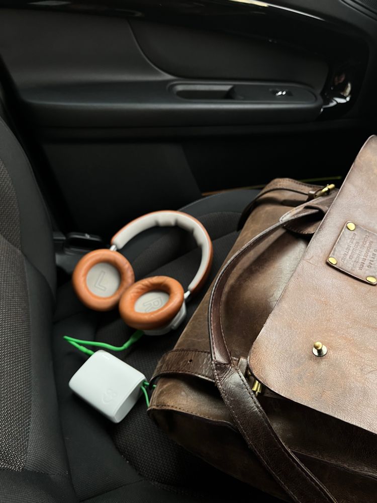 A car’s passenger seat with a brown leather bag, a pair of over-ear headphones with tan ear cushions, and an AO battery pack connected to a green charging cable. The bag has a vintage style with brass hardware, and the headphones rest beside it. The AO battery pack is small, white, and cube-shaped, lying on the seat with the charging cable extending from it. The setting suggests a moment of travel or commuting, with essential items casually placed on the seat.