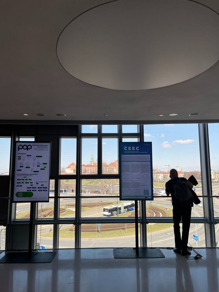 A person with short hair stands in front of large windows, silhouetted against the light, with a backpack on their back. The view outside includes a cityscape. Two digital displays are visible. The left display, titled "POP," features a flowchart or diagram with text and icons. The right display, titled "CEEC," contains text and graphs, resembling an informational poster. Outside, a road with a bus and distant buildings is visible under a partly cloudy sky.