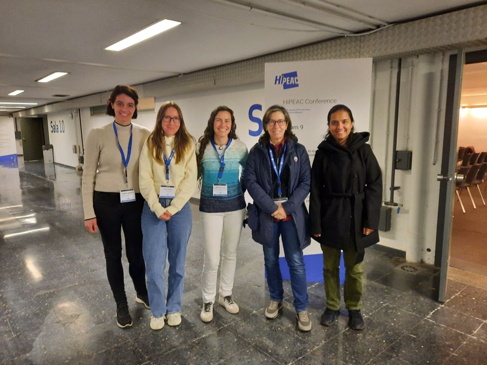 A group of five women stands in a hallway at the HiPEAC25 conference, posing for a photo. They all wear conference badges on blue lanyards. Behind them is a large sign with "HiPEAC Conference" and "Room 9" visible. The hallway has a polished floor and fluorescent lighting, with a door labeled "Sala 10" on the left. The women are dressed in casual conference attire. From left to right: the first woman has short brown hair, a light turtleneck sweater, and black pants; the second woman has long straight hair, glasses, a cream sweater, and blue jeans; the third woman has curly hair, a blue and white sweater, and white pants; the fourth woman has short hair, glasses, a dark jacket, and blue jeans; the fifth woman has long dark hair tied back, a black coat, and green pants. All are smiling.
