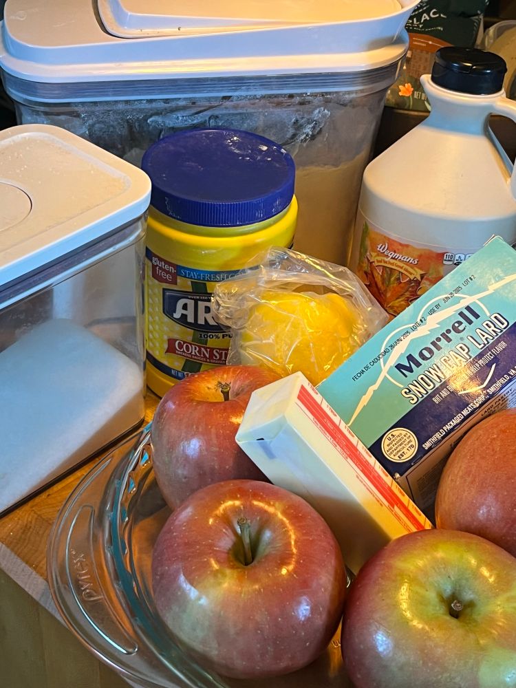 Pie ingredients: sugar in a clear plastic container, AP flour in a clear container, corn starch in a yellow plastic container, maple syrup, lemon, Macintosh apples, a stick of butter, lard & a glass pie dish on a brown cutting board 
