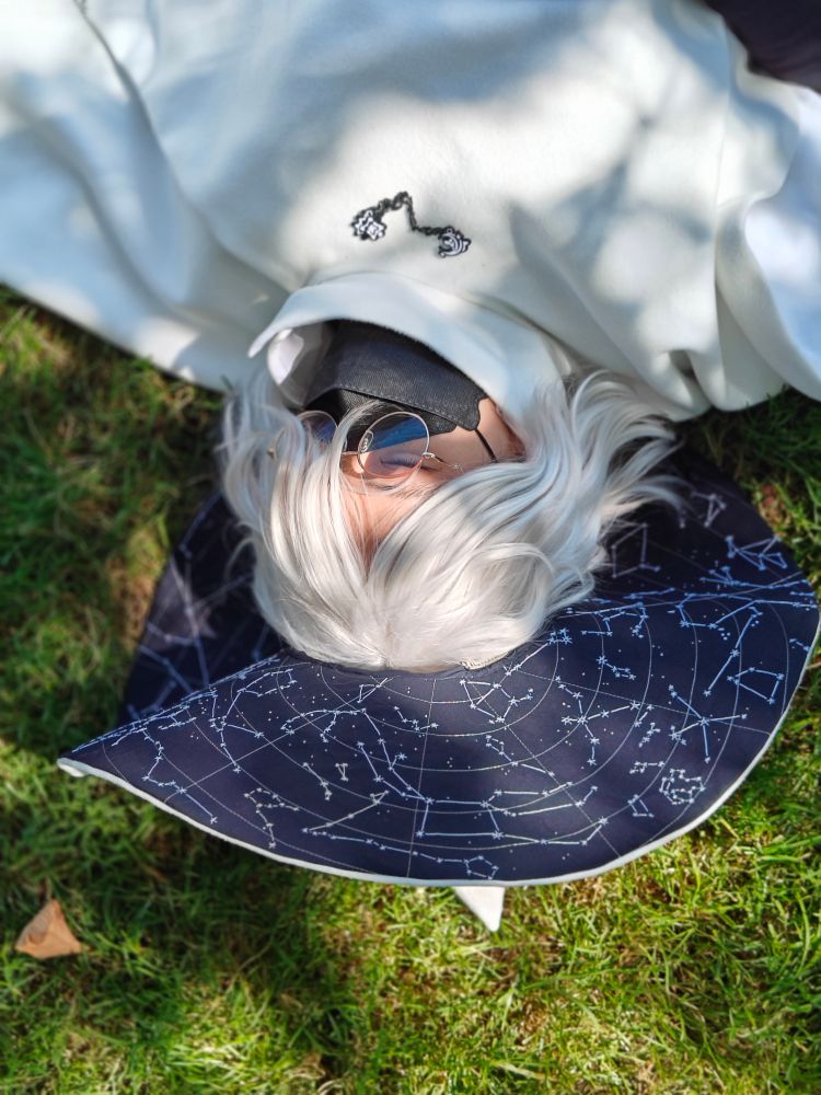 a cosplay of siffrin from in stars and time, dressed in a white cloak and hat with an embroidered star map on the underside of the brim. They're relaxing with closed eyes in dappled sunlight.