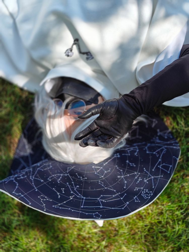 a cosplay of siffrin from in stars and time, dressed in a white cloak and hat with an embroidered star map on the underside of the brim. Their eye is open with one gloved hand outstretched, blocking the dappled sunlight in this photo.